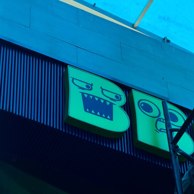 Two green dimensional B letters on a blue slatted wall, with scaffolding at the right edge.